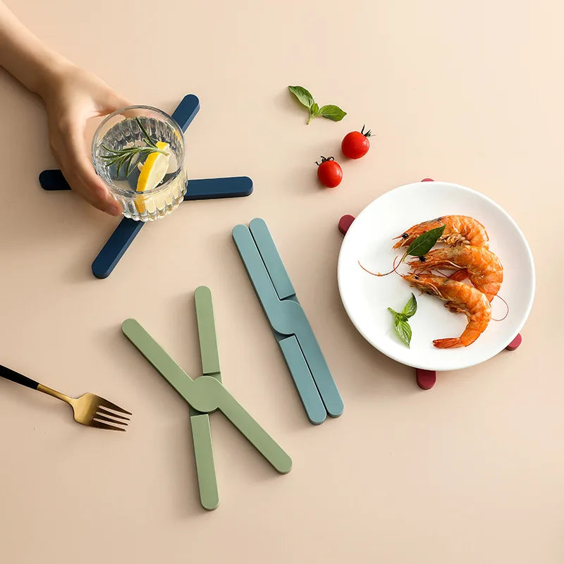 Folding cross-shaped silicone pot mats in soft colors on a kitchen table beside a plate of food